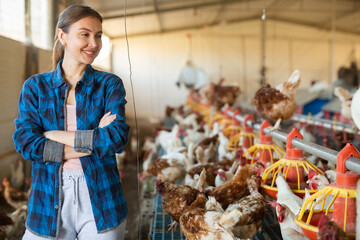Smiling female farmer in plaid shirt controling chicken feeding on farm indoor
