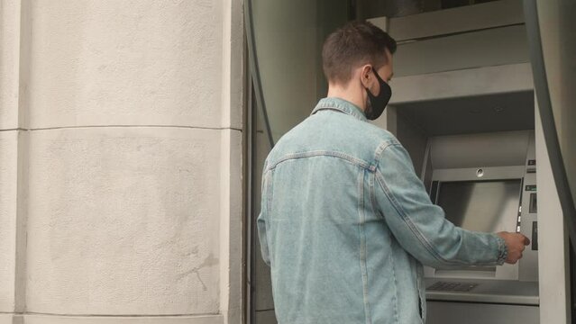 Back view portrait of young adult man wearing medical mask approaching using ATM making financial transactions. Beautiful focused caucasian male inserting card and entering pincode to withdraw cash.