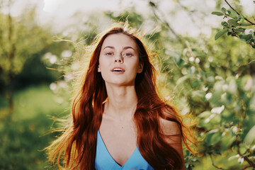 Beautiful woman with long red hair near a tree in the summer sun in the nature in the park smiling without allergies in a blue dress, the concept of health and beauty