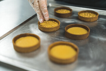 Tarts placed on a metal baking tray being decorated with golden pearls by a expert wearing a transparent glove. Blurred foreground. High quality photo