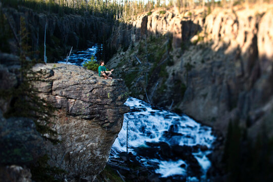 A young man meditating on the edge of a cliff looks out at the river in Montana.
