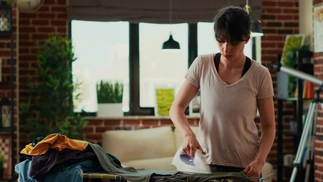 Housewife ironing shirts in living room after doing laundry, using iron with steam and heat to smooth out creases. Young adult doing household chores, weekend activity in apartment.