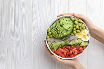 WOman holding delicious poke bowl with avocado, fish and edamame beans at white wooden table, top...