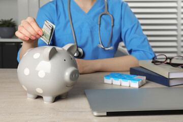 Doctor putting banknote into piggy bank at wooden table in hospital, closeup. Medical insurance