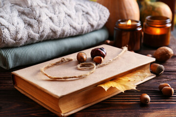 Book with autumn leaf as bookmark, scented candles and warm sweaters on wooden table