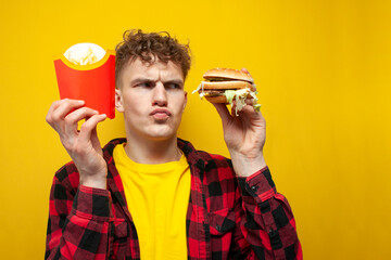 curly guy with fast food holds french fries and looks suspiciously at a burger, a man chooses...