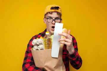 shocked consumer with grocery bag holding check and surprised by high prices on yellow background,...