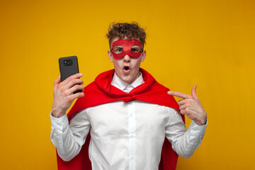 guy in a superhero costume uses a phone and shows surprise on a yellow background, super man holds a smartphone