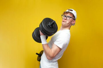 tired thin guy nerd lifts heavy dumbbells and cries on a yellow background, a confused student in glasses is training