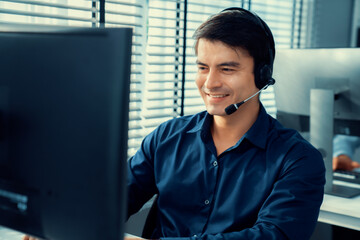 Young competent asian male call center agent working at his computer while simultaneously speaking...