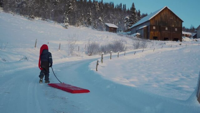 Shot of a little kid running back along the slope while having fun on the snow, enjoying sledding using red plastic shovel sled in Beitostolen, village in Innlandet county,Valdres, Beito, Norway.