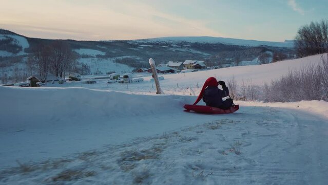 Tracking shot of a kid having fun on the snow with sledding using plastic red shovel sled in 