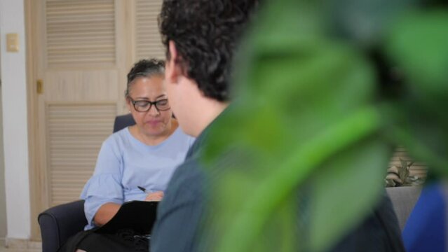 Latina Psychologist Listens to Patient and Takes Notes in Office. Young Caucasian Man Receives Therapy from Mexican Counselor Specializing in Cognitive Behavioral Therapy. 4K slow motion footage