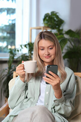 Woman with smartphone and cup of drink sitting in armchair at home