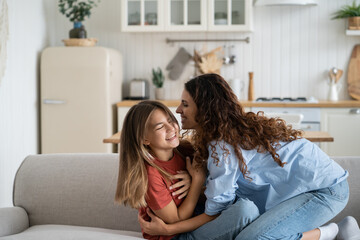 Happy caring mother hugs and tickles teenage girl to cheer up and give positive emotions during weekend or vacation. Joyful cheerful woman entertains school age daughter sits on sofa in apartment - obrazy, fototapety, plakaty