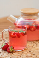 Vertical photo of a transparent glass mug with hot and fragrant strawberry tea against the background of a teapot