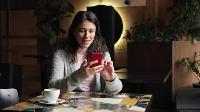 Smiling Brunette girl using smartphone sitting in coffe shop. woman texting on smartphone and laughing. Female tapping on mobile phone. View photos and videos on the phone.