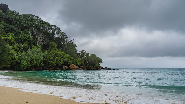 A secluded beach on a tropical island. The waves of the turquoise ocean are foaming on the sand. Boulders near the water. A hill overgrown with tropical vegetation, against a cloudy sky. Seychelles. 