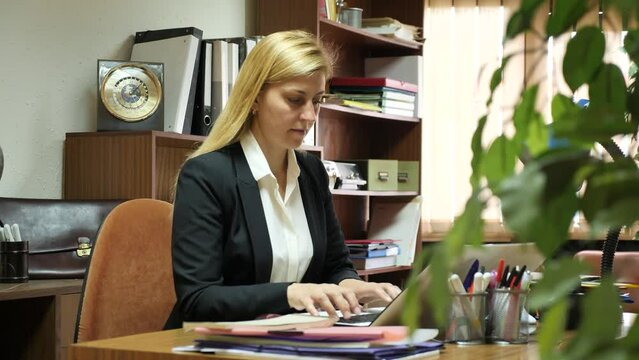 Middle age european businesswoman in business suit sitting in office at workplace and working at laptop