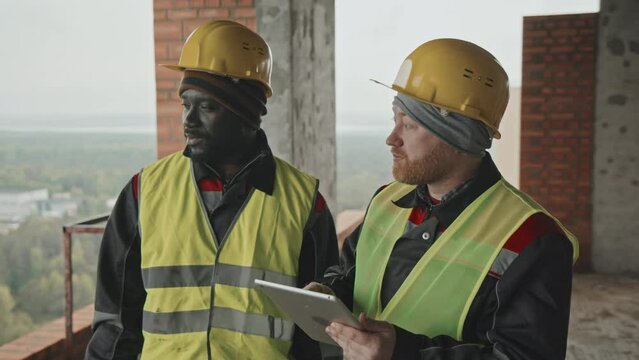 Ethnically diverse construction worker and foreman wearing uniform looking at plan on digital tablet screen and discussing issues