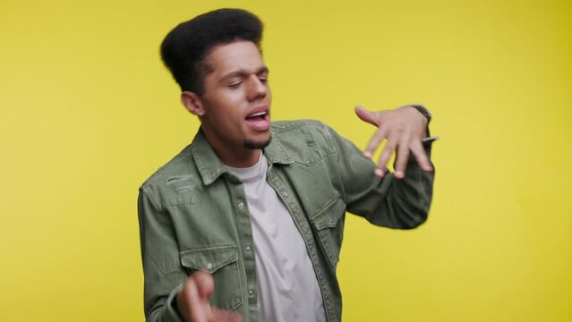 Funny smiling African American young guy 20s in casual clothes on yellow background in studio. People lifestyle concept. Black man dancing fooling around having fun expressive gesticulating hands.