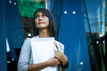 Young Asian executive woman holding work documents outside glass office building. Pensive Thai transgender person with files in hand and doubtful look thinking about project ideas