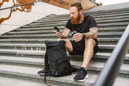 Young handsome bearded tattooed redhead man with bottle and phone