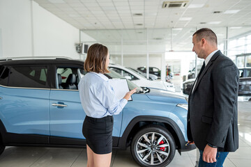 young manager in suit holding clipboard with female client  talking to sales modern car