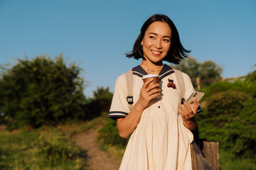 Young smiling asian woman holding coffee and mobile phone while walking in green park