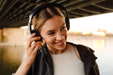 Young smiling woman listening music with headphones while standing on embankment