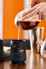 Shot of female hands pouring tea into a dark night ceramic color printed cup in chinese style. The raised bowl with an image of flying cranes is located on the wooden table on the orange background.