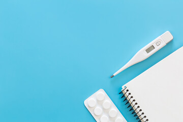 Electronic thermometer, pills and notepad on a blue background, flat lay.
