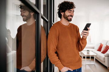 Smiling young man using mobile phone standing by glass door at home