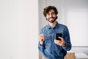 Happy young entrepreneur with smart phone standing by window in office
