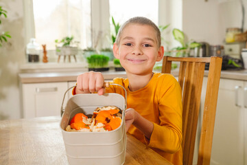 Smiling boy showing mandarin peels in compost bucket at dining table