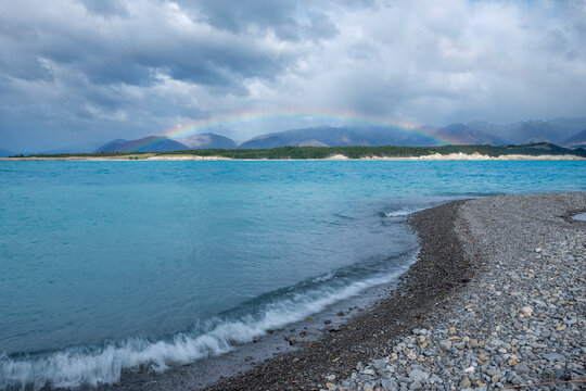 New Zealand, South Island, Rocky shore ofLake Pukaki with rainbow arching in background