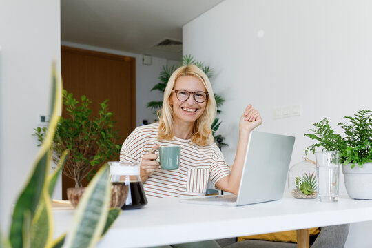 Happy freelancer with tea cup and laptop sitting at desk - Powered by Adobe