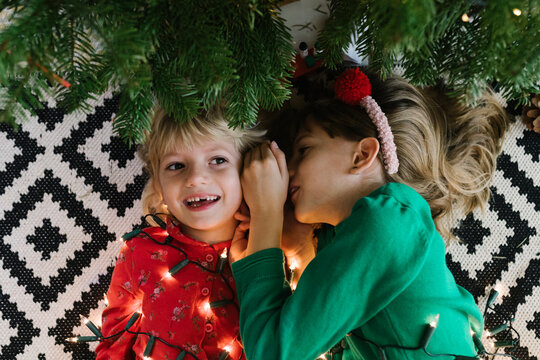 Girl whispering in sister's ear lying down with illuminated string lights near Christmas tree