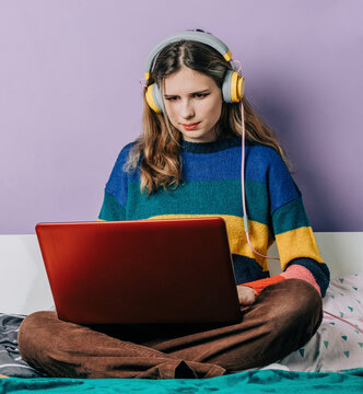 Girl wearing headphones doing home work using laptop in front of purple wall