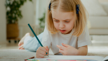 Cute concentrated Caucasian little kid girl draw color pencil lying on floor at home play alone....
