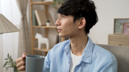 Fototapeta premium cheerful asian japanese young male looking out of window and enjoying the tranquil morning and coffee from cup in the living room at home
