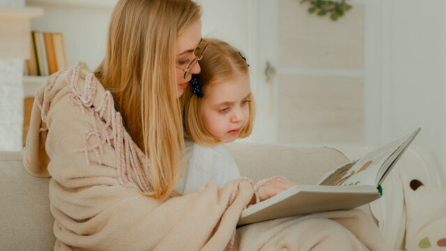 Little small Caucasian daughter child girl kid baby with mother in living room reading literature looking at photo album mom read book showing pictures explain at couch sofa family elementary learning