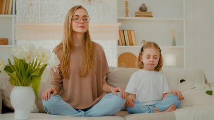 Caucasian woman mom with little child girl sitting home at couch in lotus position closed eyes meditate together. Family meditating mother teach meditation small daughter adorable baby kid breath