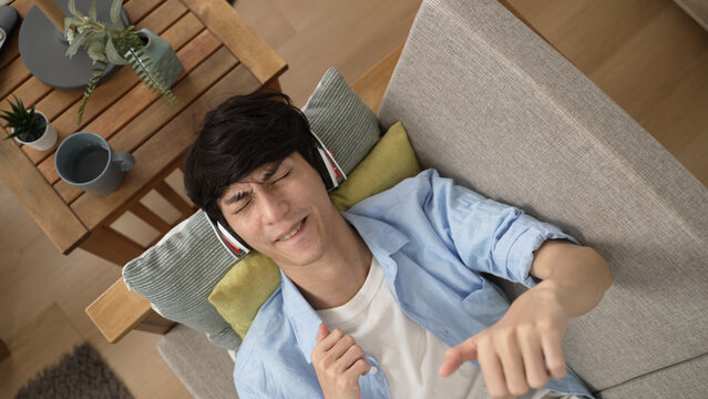 Top View With Zoom Out Cheerful Asian Chinese Guy Looking Upward And Singing With Emotional Hand Gesture While Lying On Couch Enjoying Song From Headset At Home.
