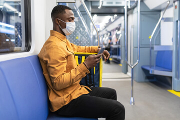 An African-American courier in a protective mask holds a mobile phone while riding in a subway car with a yellow backpack