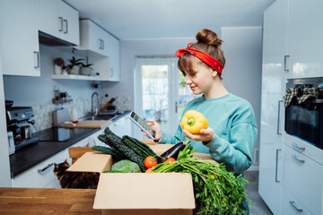 Online home food delivery. Woman checking her online order list on her phone. Cardboard box with fresh vegetables and fruits standing on the kitchen table. Local farmer food. Start of a healthy life.