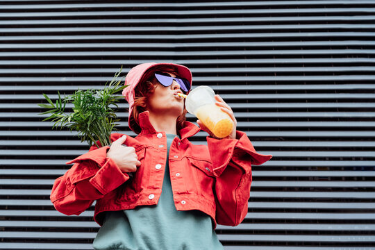 Hipster fashion woman in bright clothes, heart shaped glasses, bucket hat drinking fruity sugar flavored tapioca pearl bubble tea and holding green potted plant on the gray striped wall background.