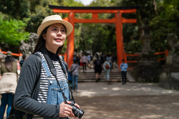 cheerful Asian Chinese girl traveler carrying camera and looking into distance while admiring beautiful view near a scarlet torii gate of kasuga grand shrine in nara japan