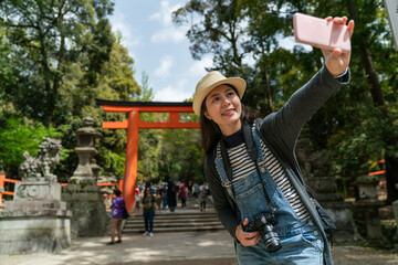 cheerful Asian Japanese girl using mobile phone to take selfie picture with scarlet torii gate at background while traveling to kasuga grand shrine in nara japan on a clear sunny day