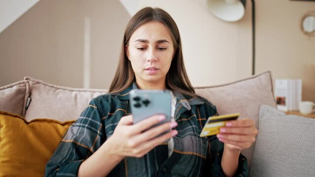 Young woman sits on couch at home and enters bank card details for payment in online store using mobile phone. Female smiles and enjoys shopping. Buy an expensive dream item. Gift, success, wealth.
